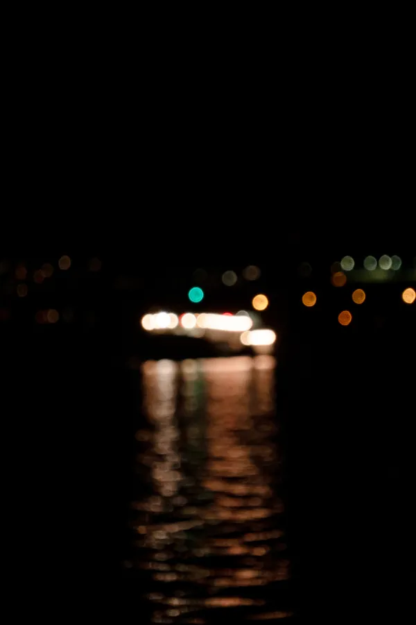 A ferry crosses the lake at night, its lights blurred into soft circles of white, orange, and teal against the dark water.
