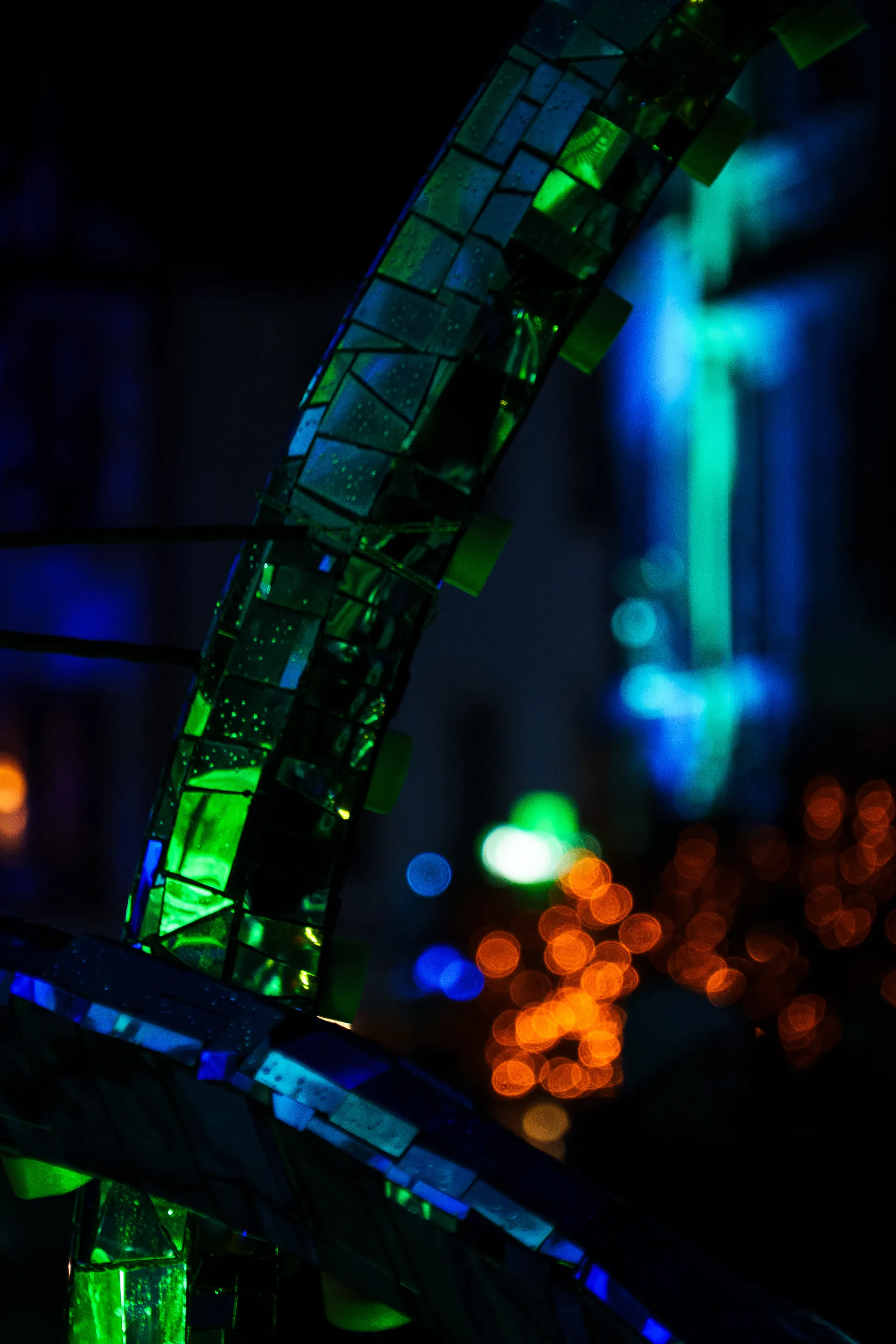 A mirrored structure reflects vibrant green and blue light against the night, creating an abstract kaleidoscope of colors with warm bokeh lights below.