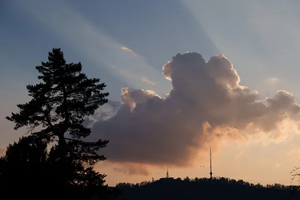 A towering cumulonimbus cloud lit by the setting sun with the Uetliberg tower silhouetted on the ridge below.