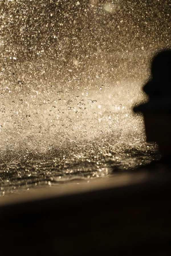 A silhouette emerges against a cascade of golden water droplets backlit by the sun.