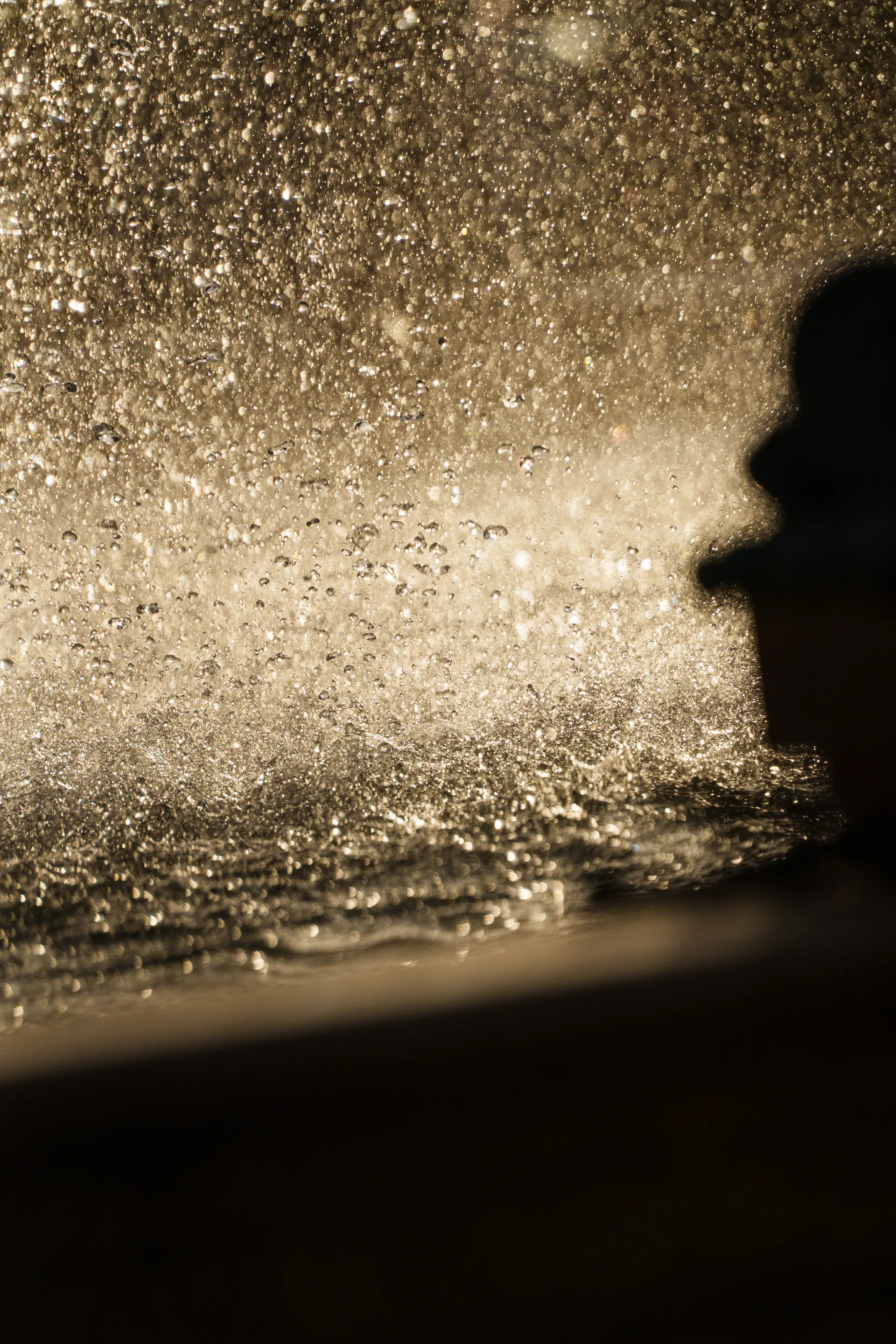 A silhouette emerges against a cascade of golden water droplets backlit by the sun.