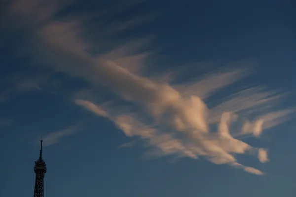The Eiffel Tower sits small in the corner while dramatic wispy cirrus clouds sweep across a deep blue sky.