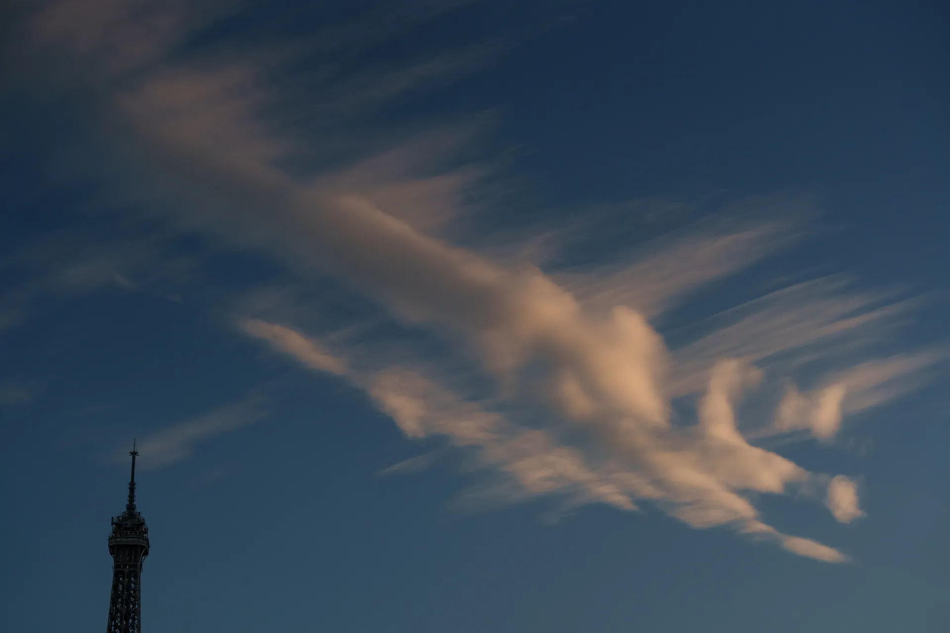 The Eiffel Tower sits small in the corner while dramatic wispy cirrus clouds sweep across a deep blue sky.