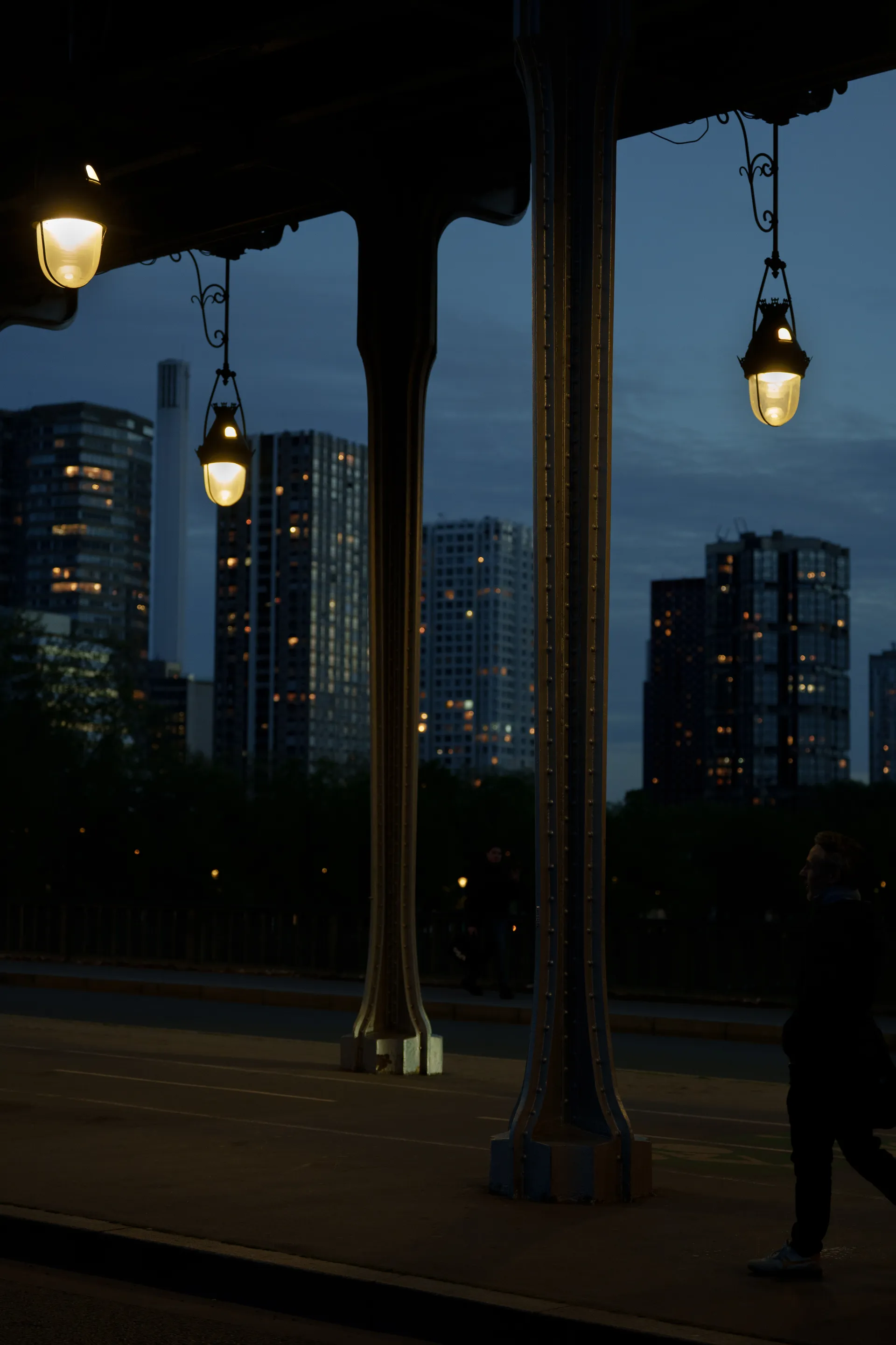 Ornate lanterns hang beneath an iron bridge at dusk, framing a distant city skyline lit from within.