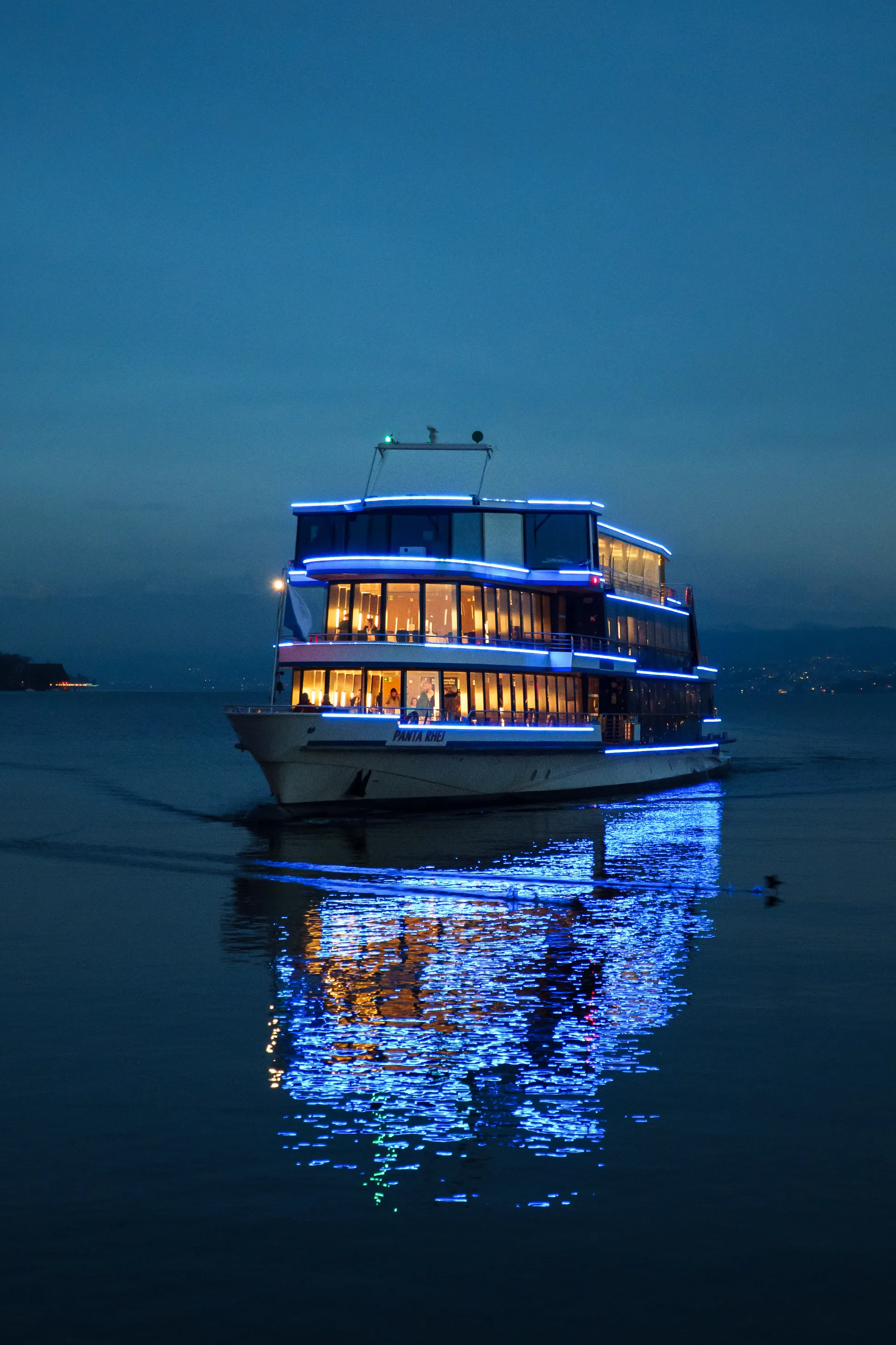A passenger boat illuminated with blue LED lights glides across calm water at dusk, warm interior lights glowing through windows as reflections dance on the surface.