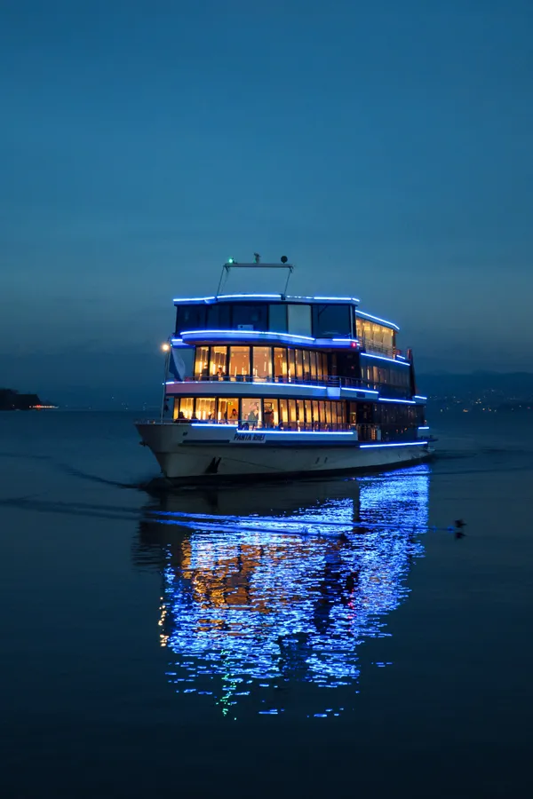 A passenger boat illuminated with blue LED lights glides across calm water at dusk, warm interior lights glowing through windows as reflections dance on the surface.