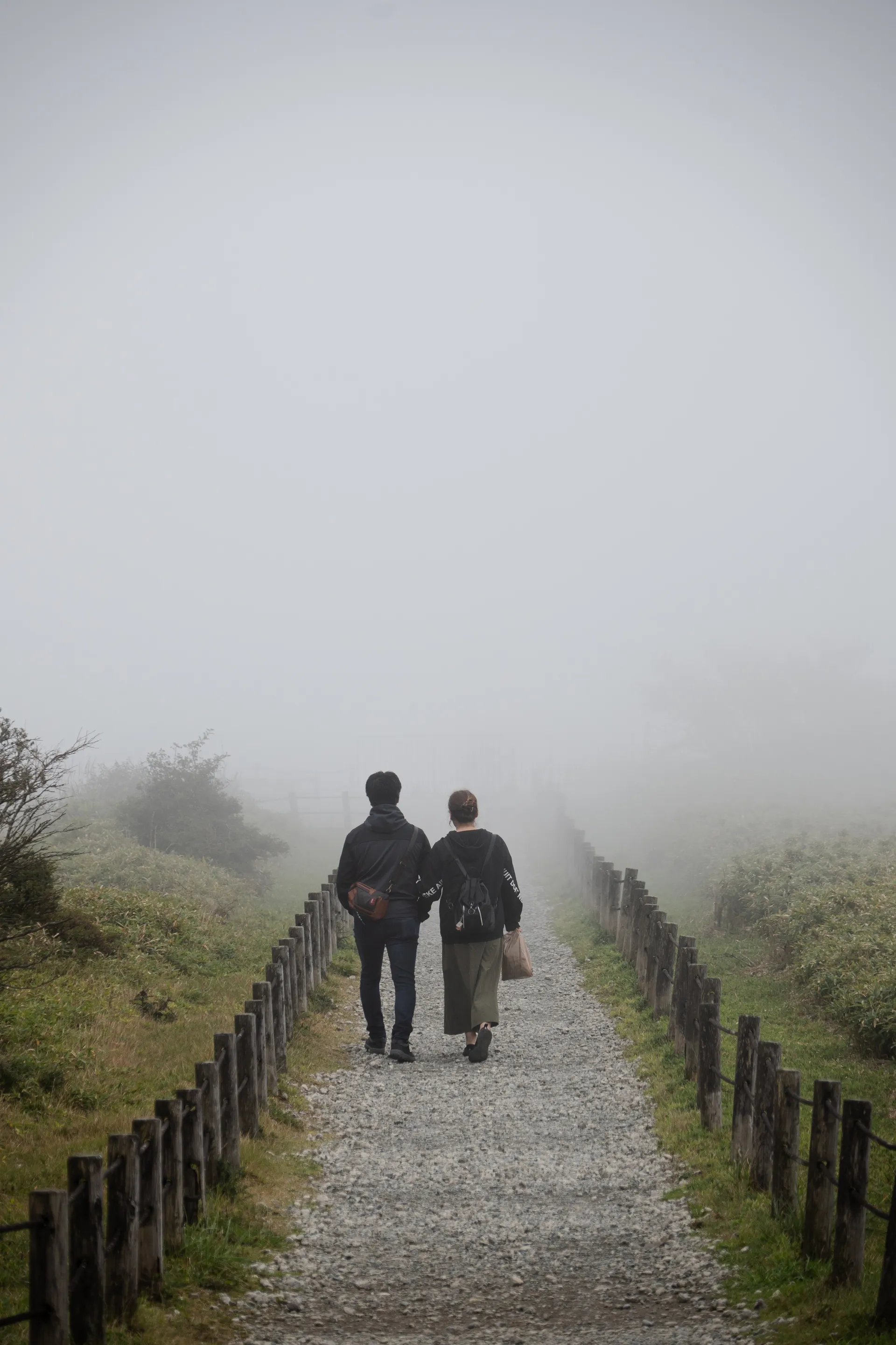 Two hikers walk along a gravel path lined with wooden posts, disappearing into the mountain mist.