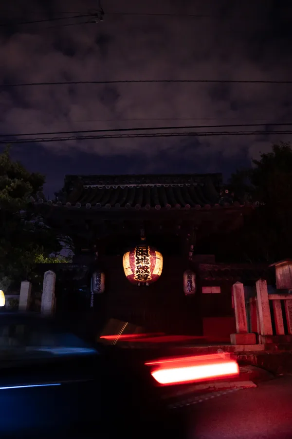 A glowing paper lantern illuminates an ancient temple gate as car lights streak past.