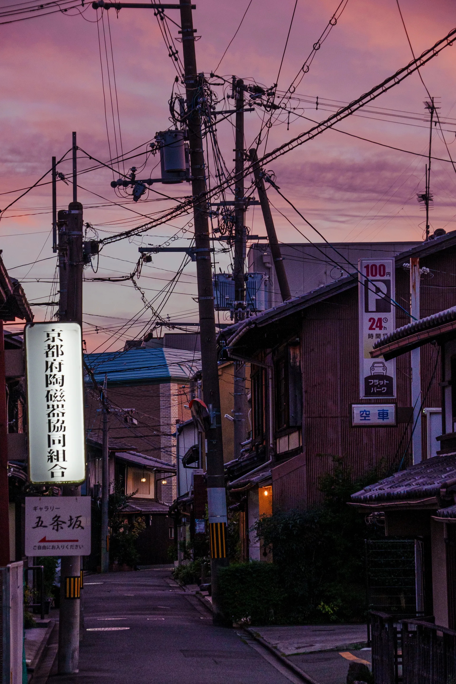 A narrow Kyoto street lined with traditional buildings and tangled power lines glows under a pink and purple dusk sky.