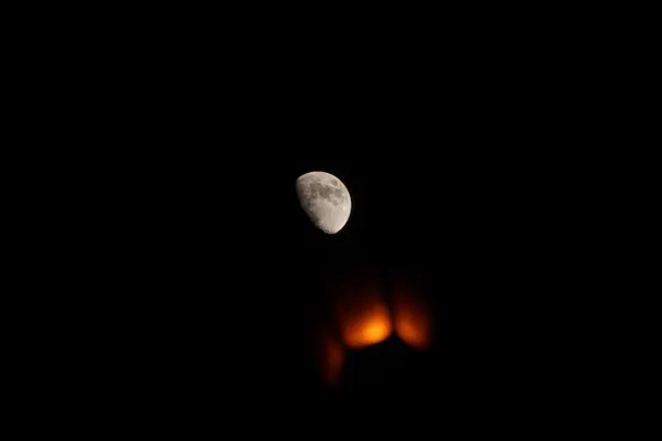 A waxing moon hangs sharp and bright above a warm orange glow emerging from the darkness below.
