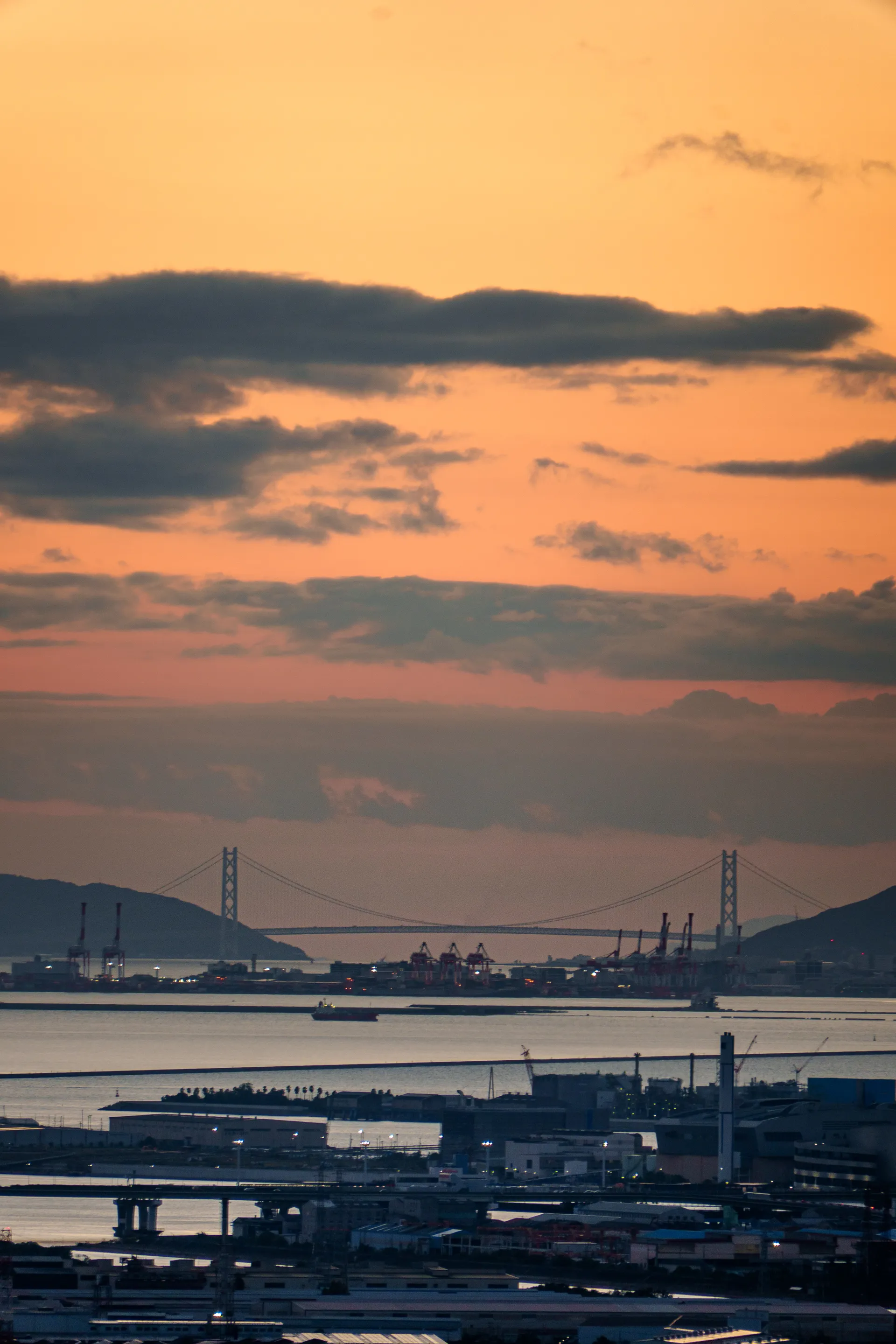 A suspension bridge stretches across a harbor at sunset, framed by dramatic orange and gray clouds, with industrial port infrastructure in the foreground.