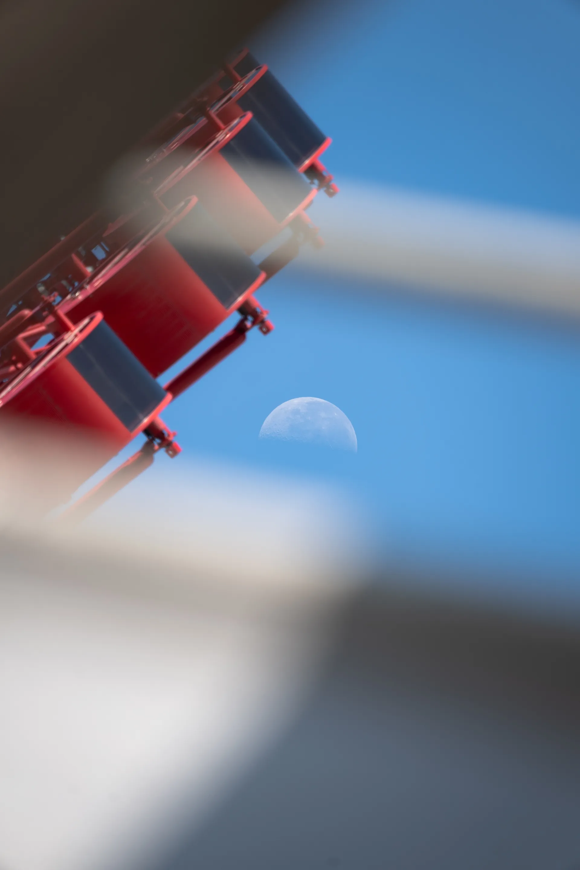 A red ferris wheel against a clear blue sky, while a delicate crescent moon hangs sharp and still in the background.