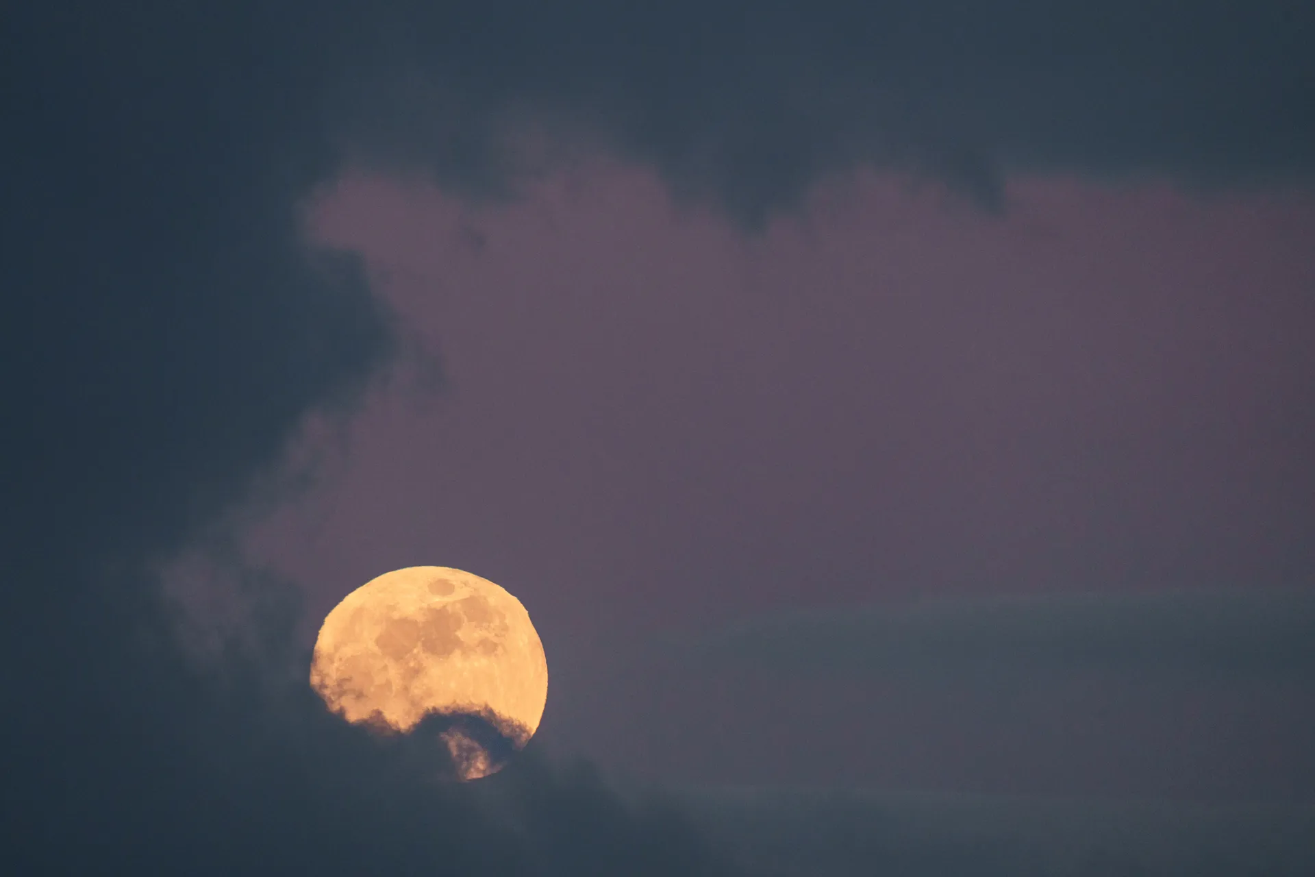 A golden full moon rises behind dark clouds, its warm glow contrasting with the blue-purple dusk sky.