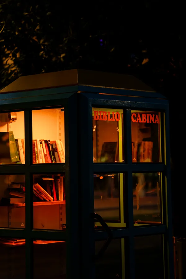 A small phone-booth-style free library glows warm orange against the night, books lining its illuminated shelves behind glass.
