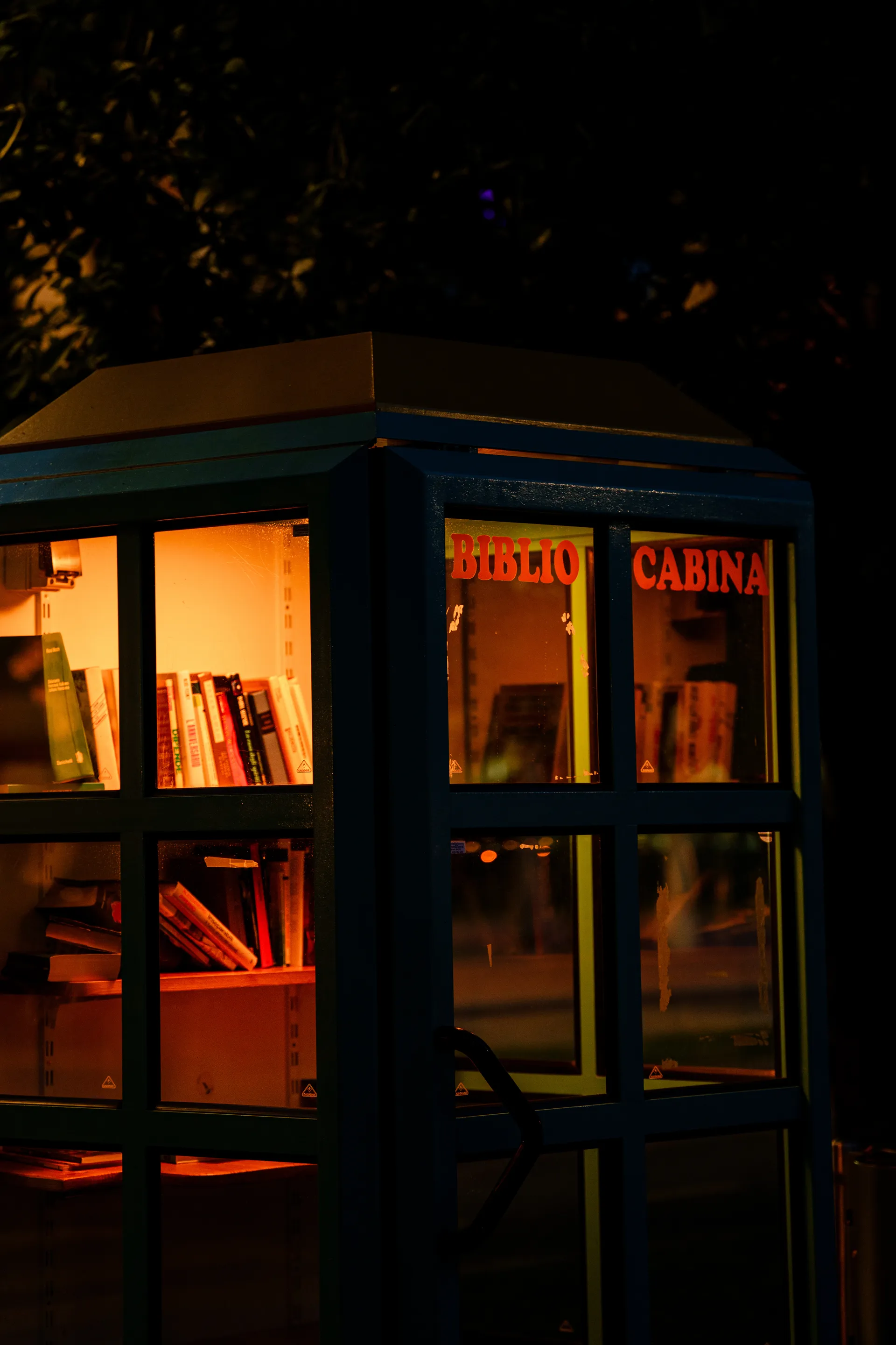 A small phone-booth-style free library glows warm orange against the night, books lining its illuminated shelves behind glass.
