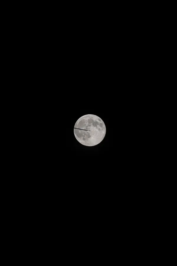 An airplane crosses the face of the full moon, a fleeting silhouette against the lunar surface.