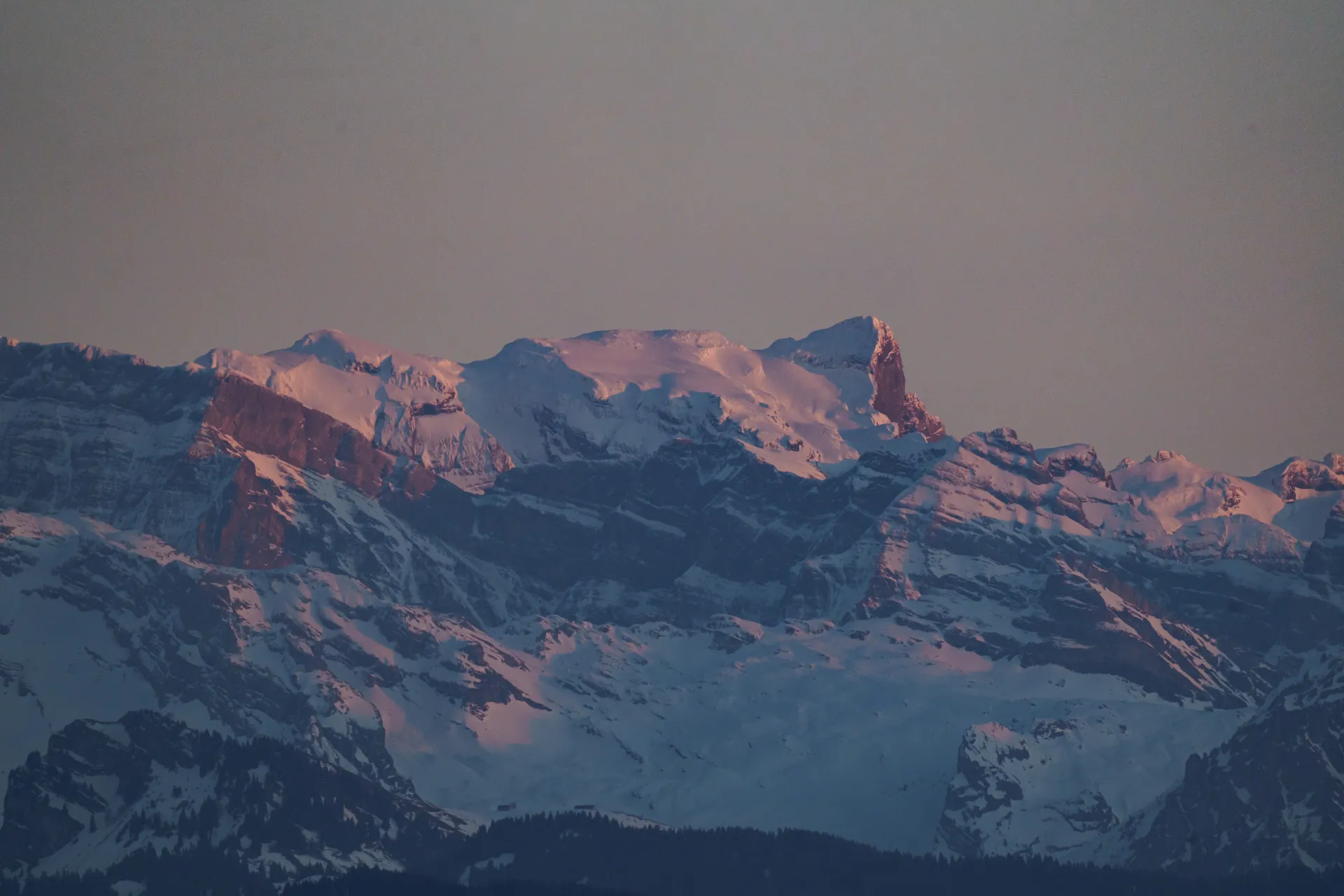 Snow-covered Alpine peaks catch the last warm light of the day, their ridges glowing pink and orange against a muted sky.