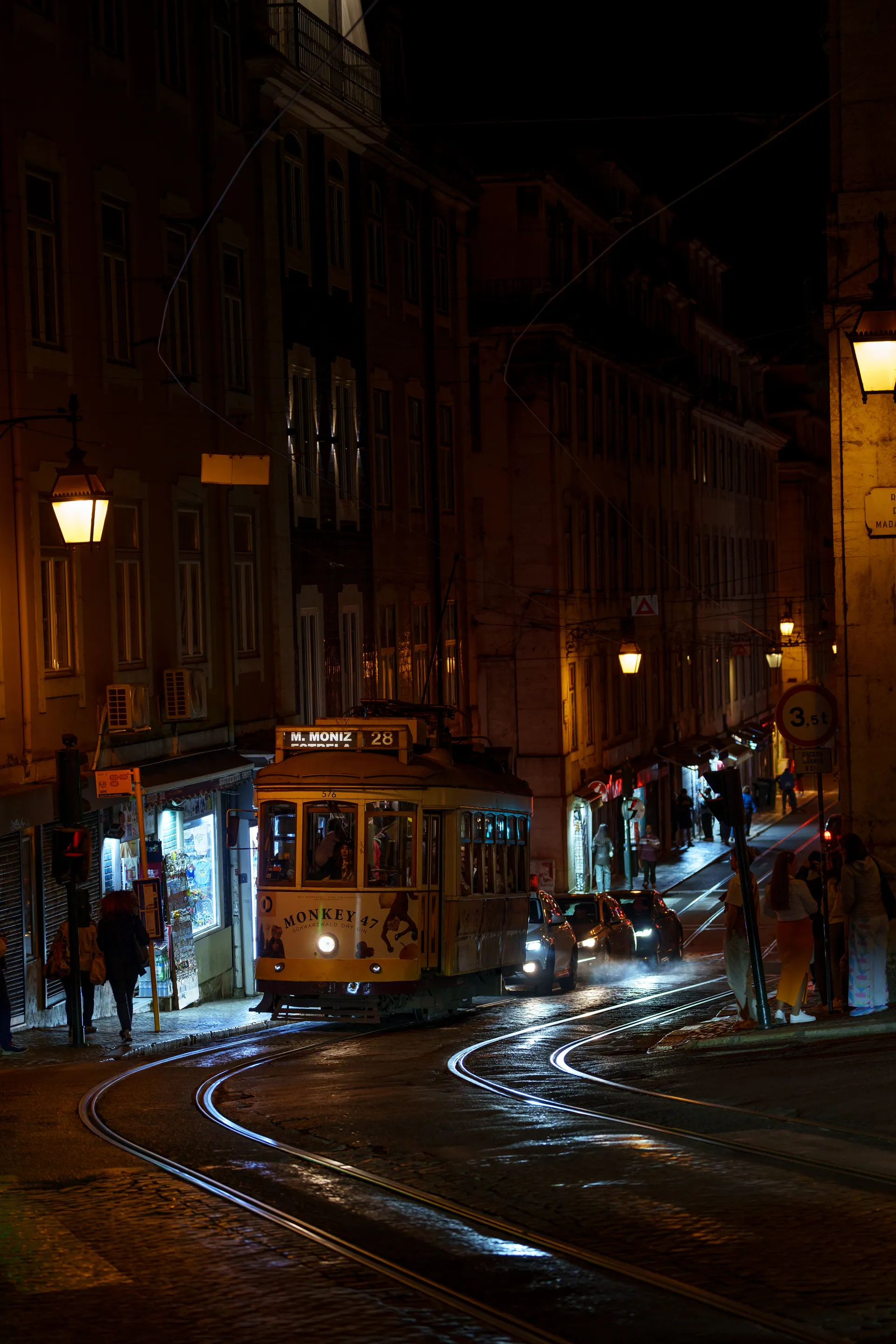 A yellow Lisbon tram waits on curved cobblestone tracks at night, warm streetlamps glowing along the narrow street.