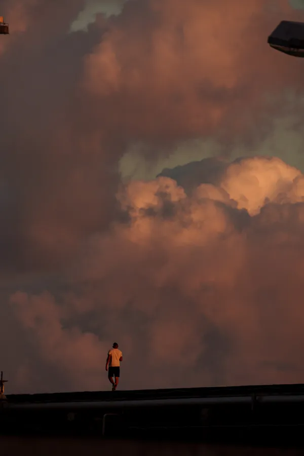 A lone figure walks along an elevated edge, silhouetted against towering sunset clouds glowing in warm orange and pink tones.