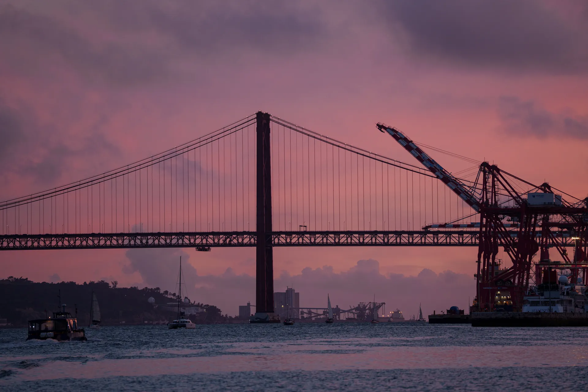 The Ponte 25 de Abril stretches across the Tagus at dusk, port cranes silhouetted on the far bank under pink and purple clouds.