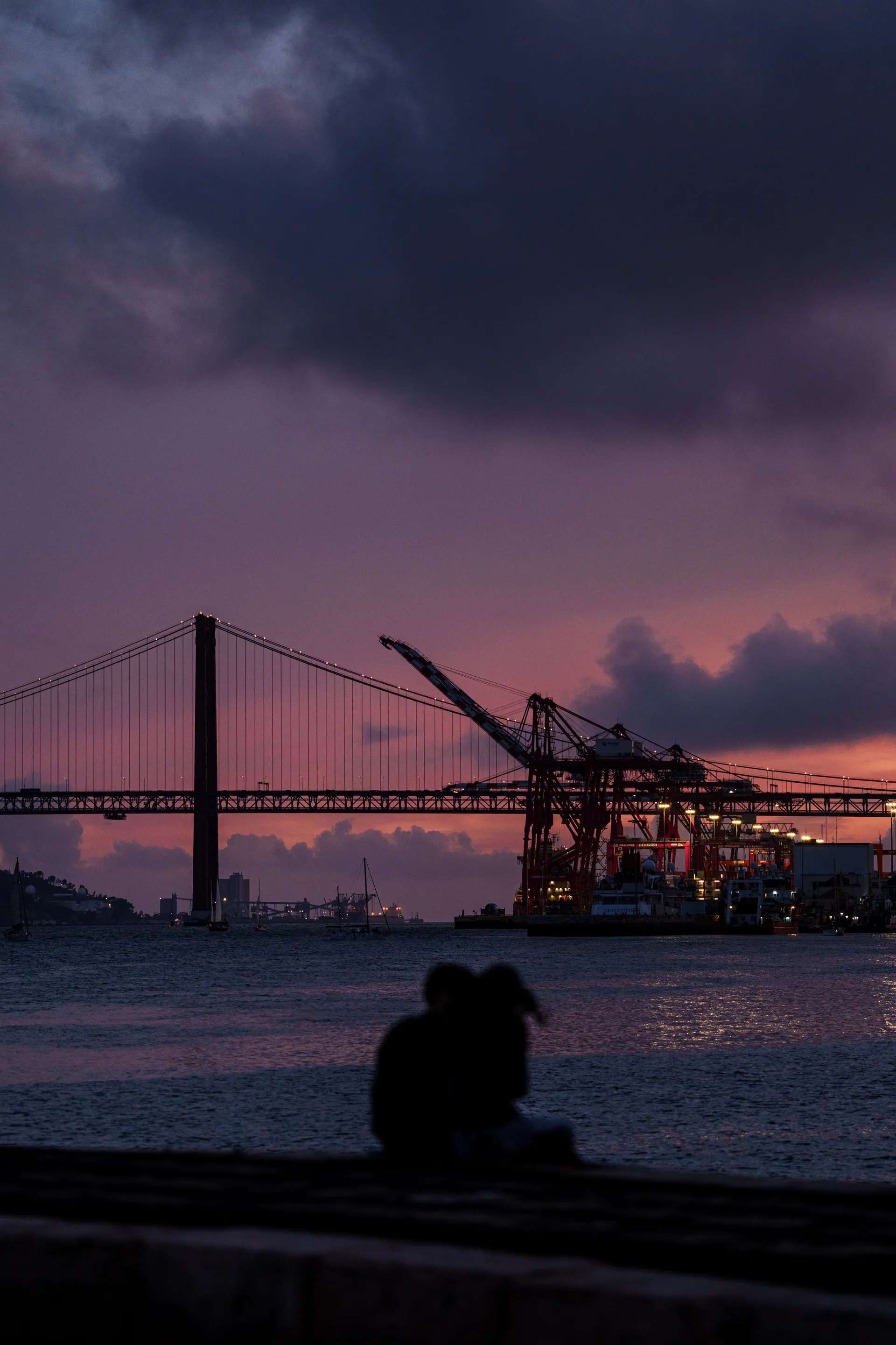 A silhouetted couple sits embracing at the waterfront, the Ponte 25 de Abril and port cranes rising behind them under a deep purple dusk sky.