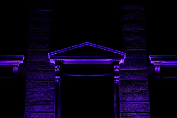 A neoclassical entrance in Neuchâtel glows under deep violet uplighting, its pediment and columns outlined against total darkness.