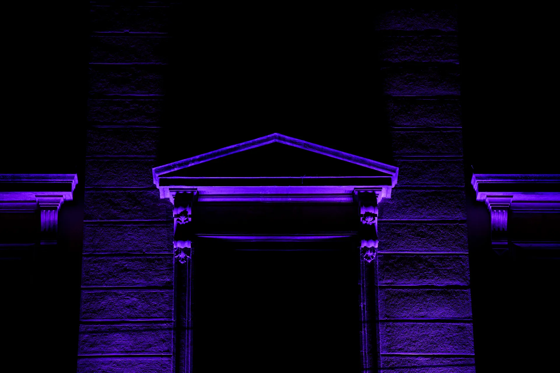 A neoclassical entrance in Neuchâtel glows under deep violet uplighting, its pediment and columns outlined against total darkness.