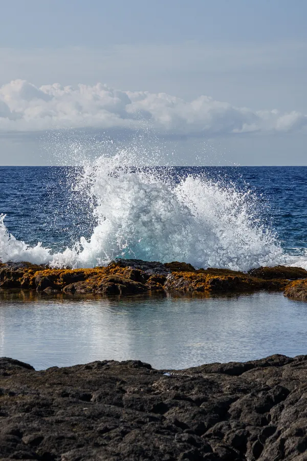 A powerful wave explodes against the volcanic rocks, sending spray into the tropical air.
