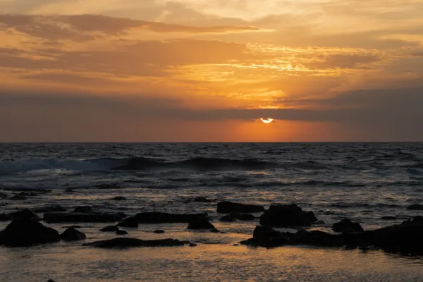 The sun dips below the horizon, painting the sky in golden hues over a rocky shoreline.