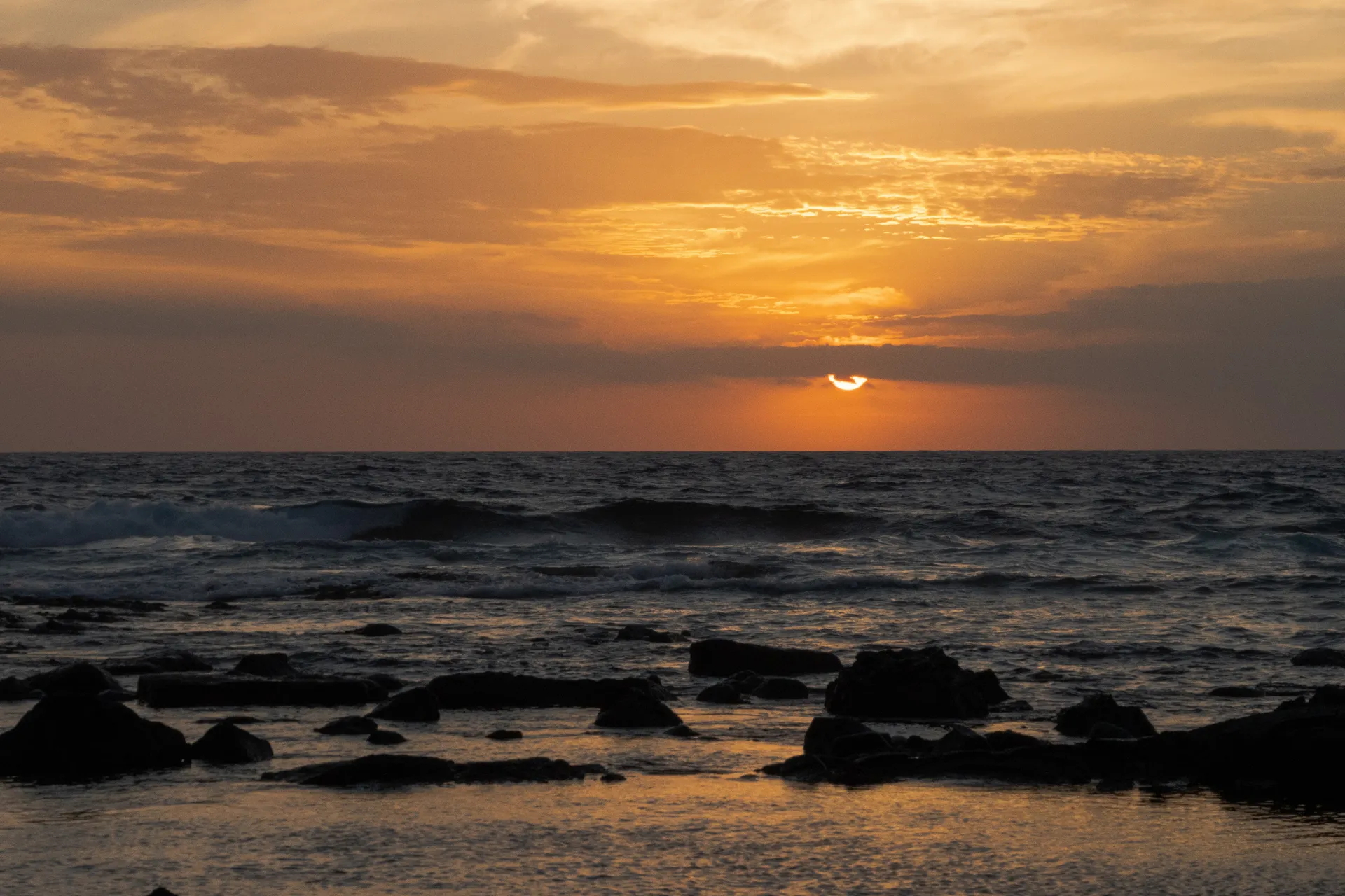 The sun dips below the horizon, painting the sky in golden hues over a rocky shoreline.