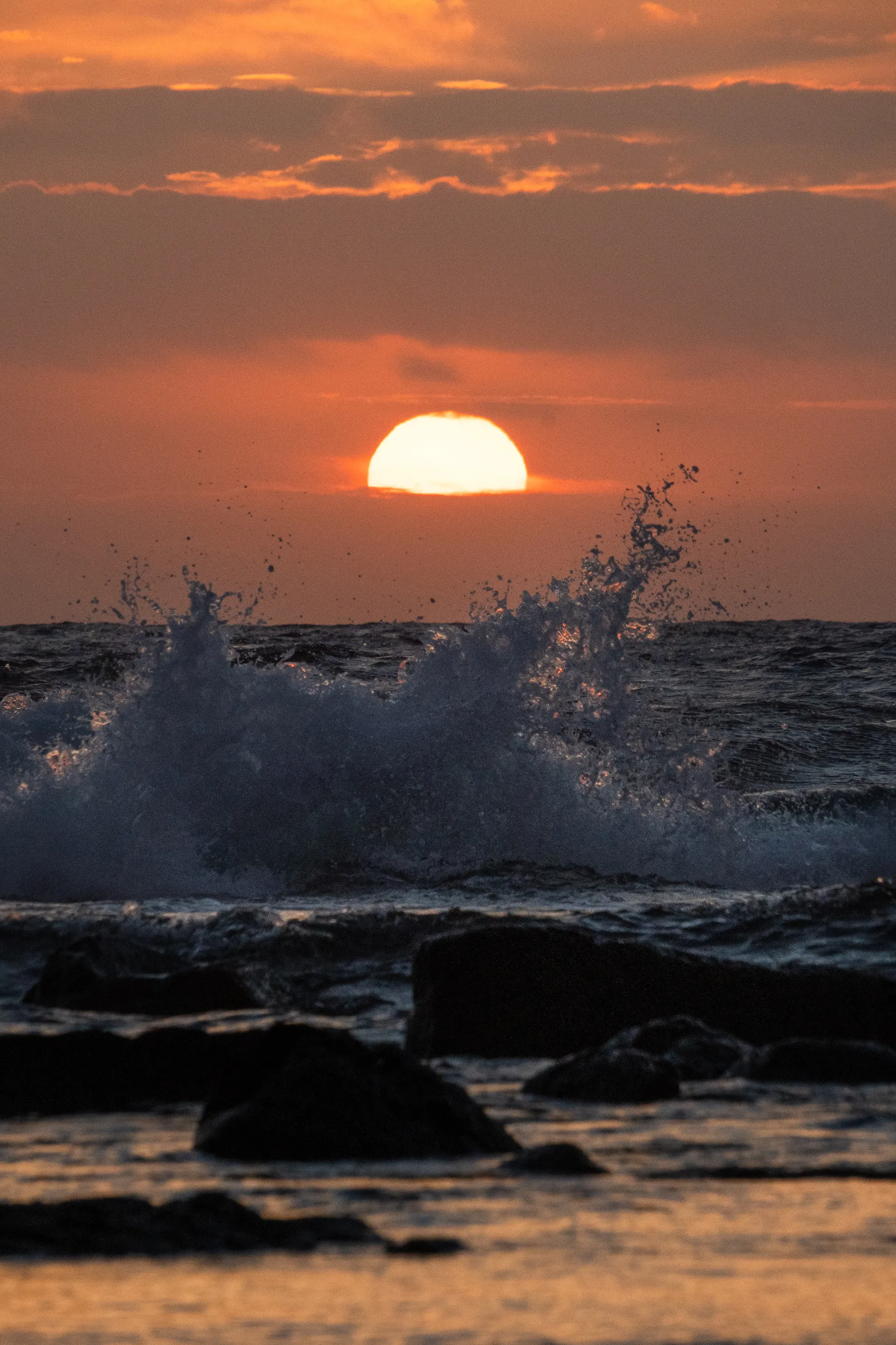 A wave crashes against dark volcanic rocks as the sun sinks into the ocean horizon, casting an orange glow across the water.