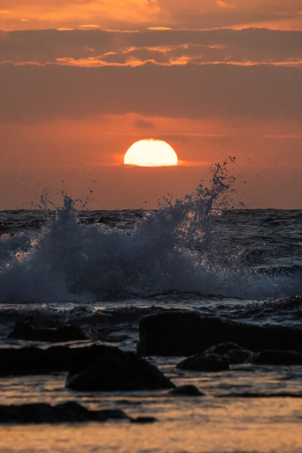 A wave crashes against dark volcanic rocks as the sun sinks into the ocean horizon, casting an orange glow across the water.