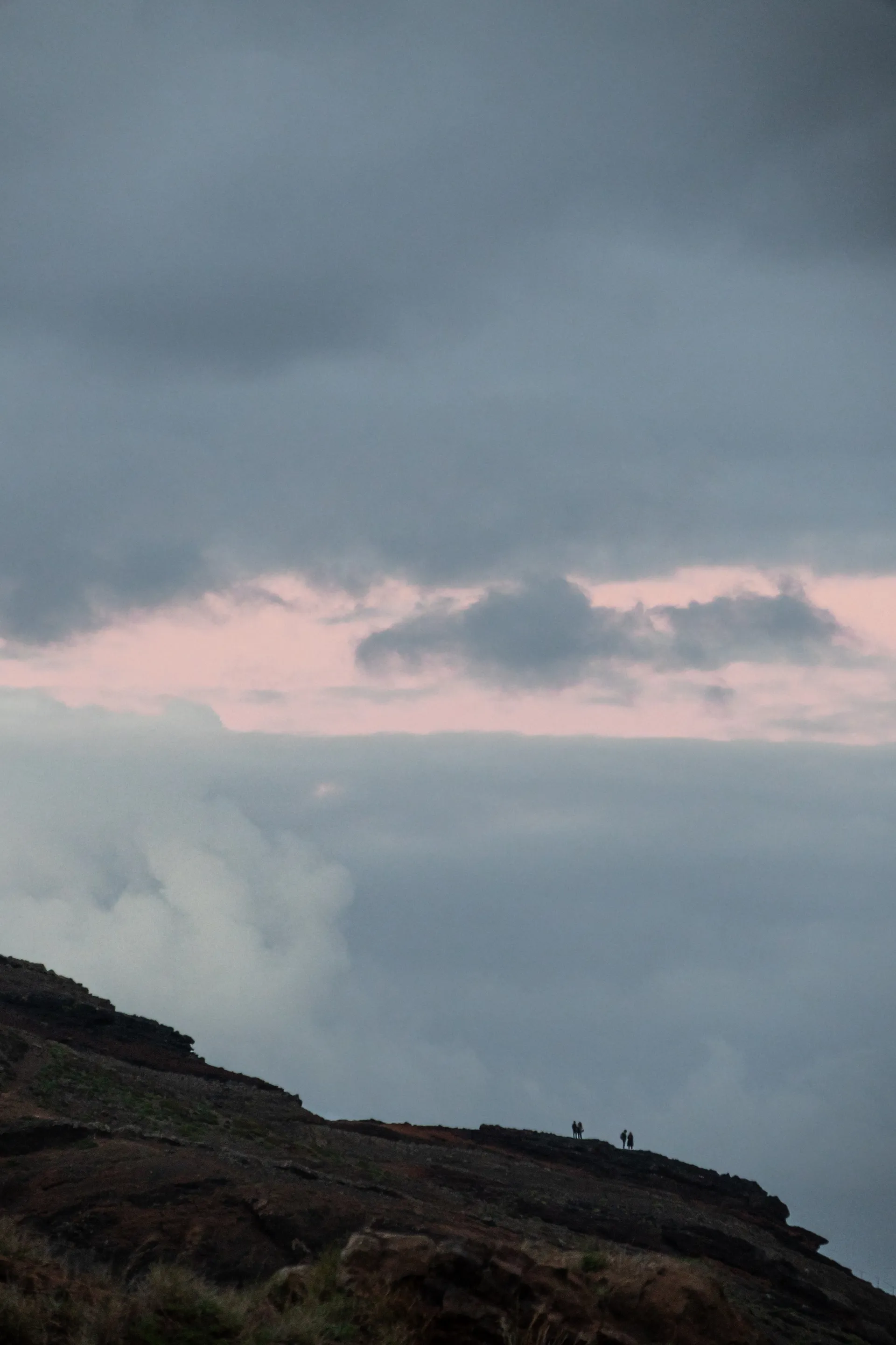Silhouettes of two couples stand on a cliff edge beneath dramatic clouds tinged with pink twilight.