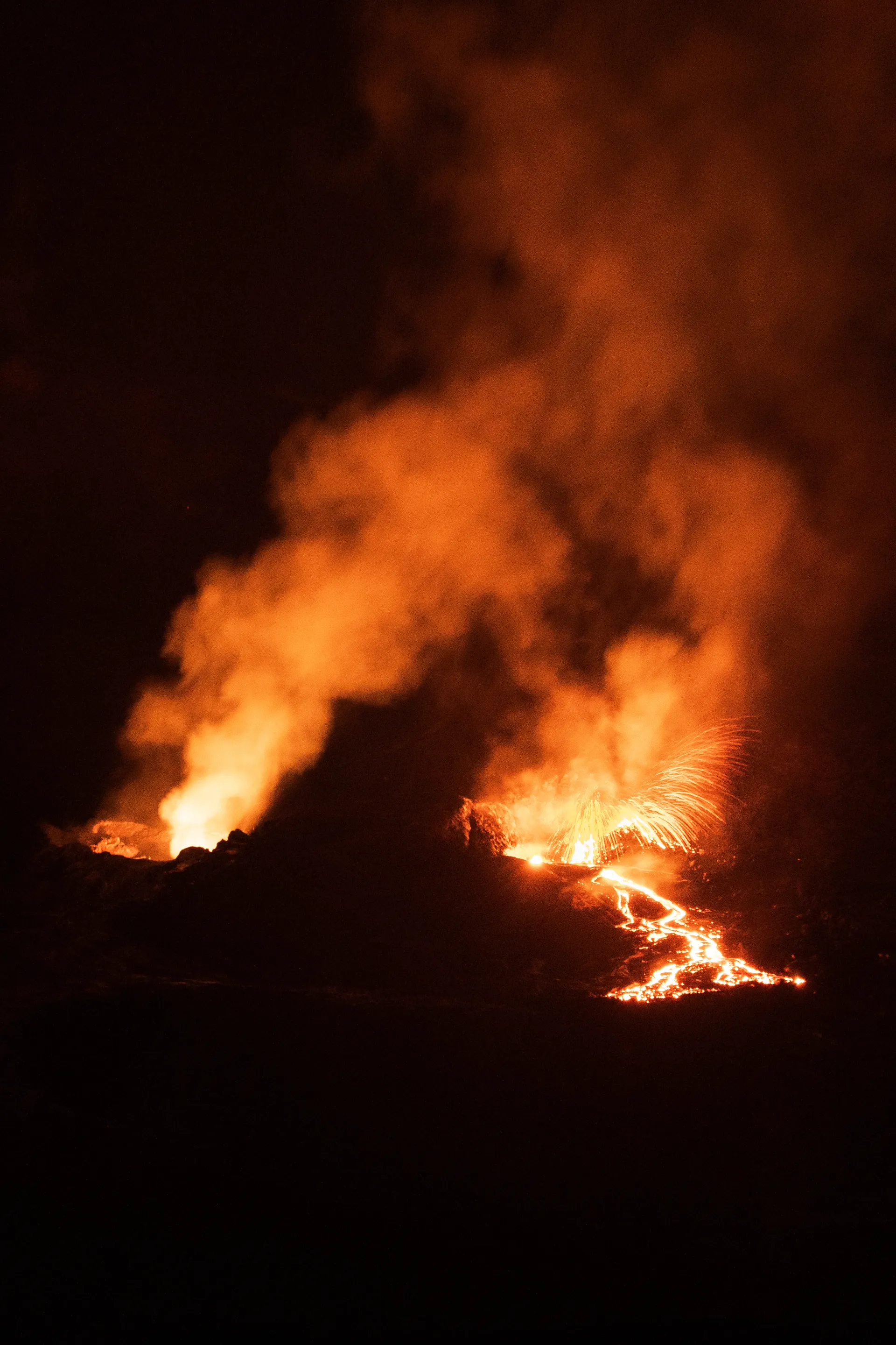 Molten lava from Kīlauea flows across dark volcanic rock, sparks flying into the night sky.