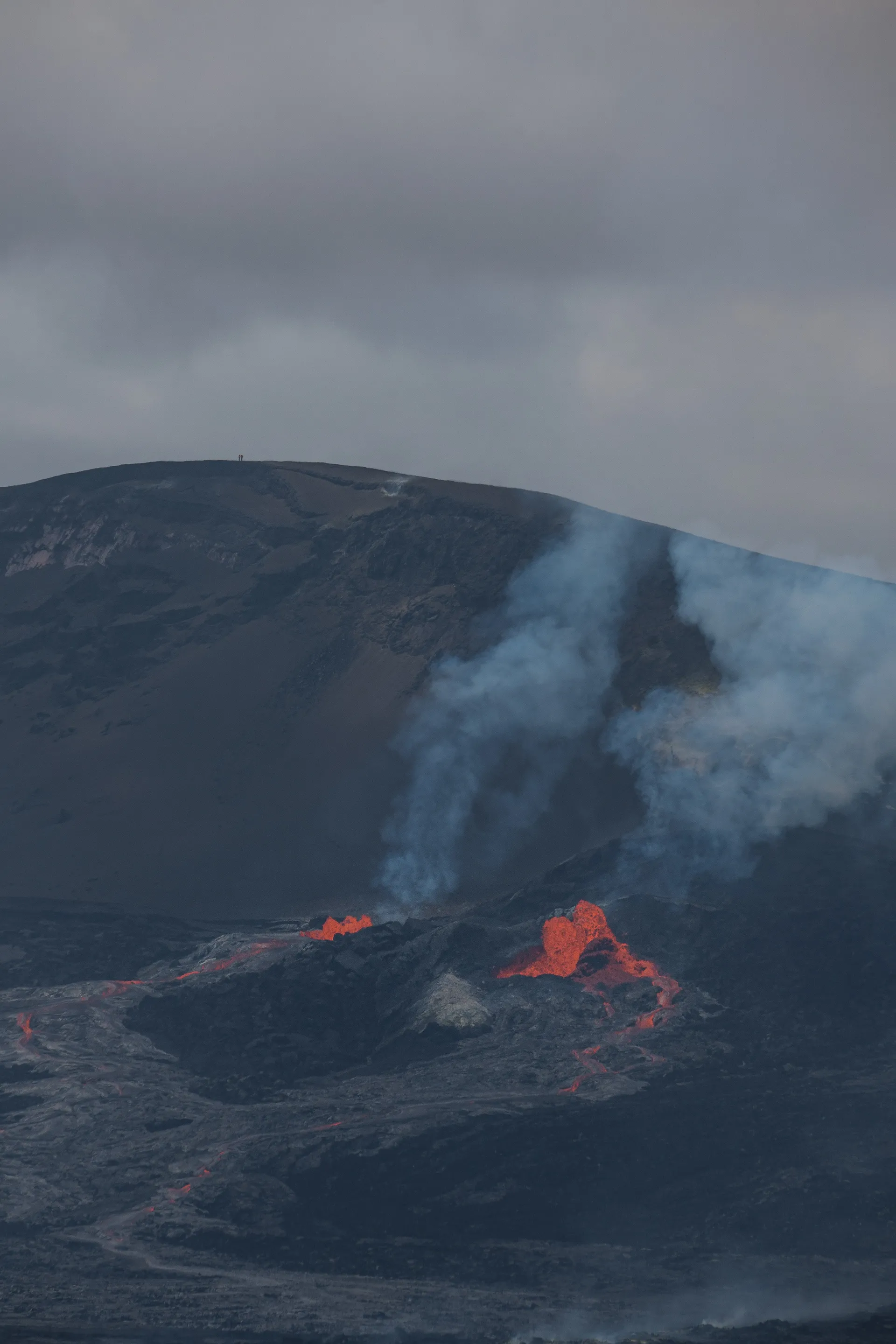 Molten lava glows bright orange against the dark volcanic landscape, smoke rising into the overcast sky.