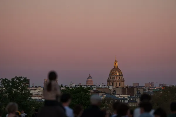 The golden dome of Les Invalides glows in pink dusk light above the Paris skyline, watched by silhouetted onlookers.