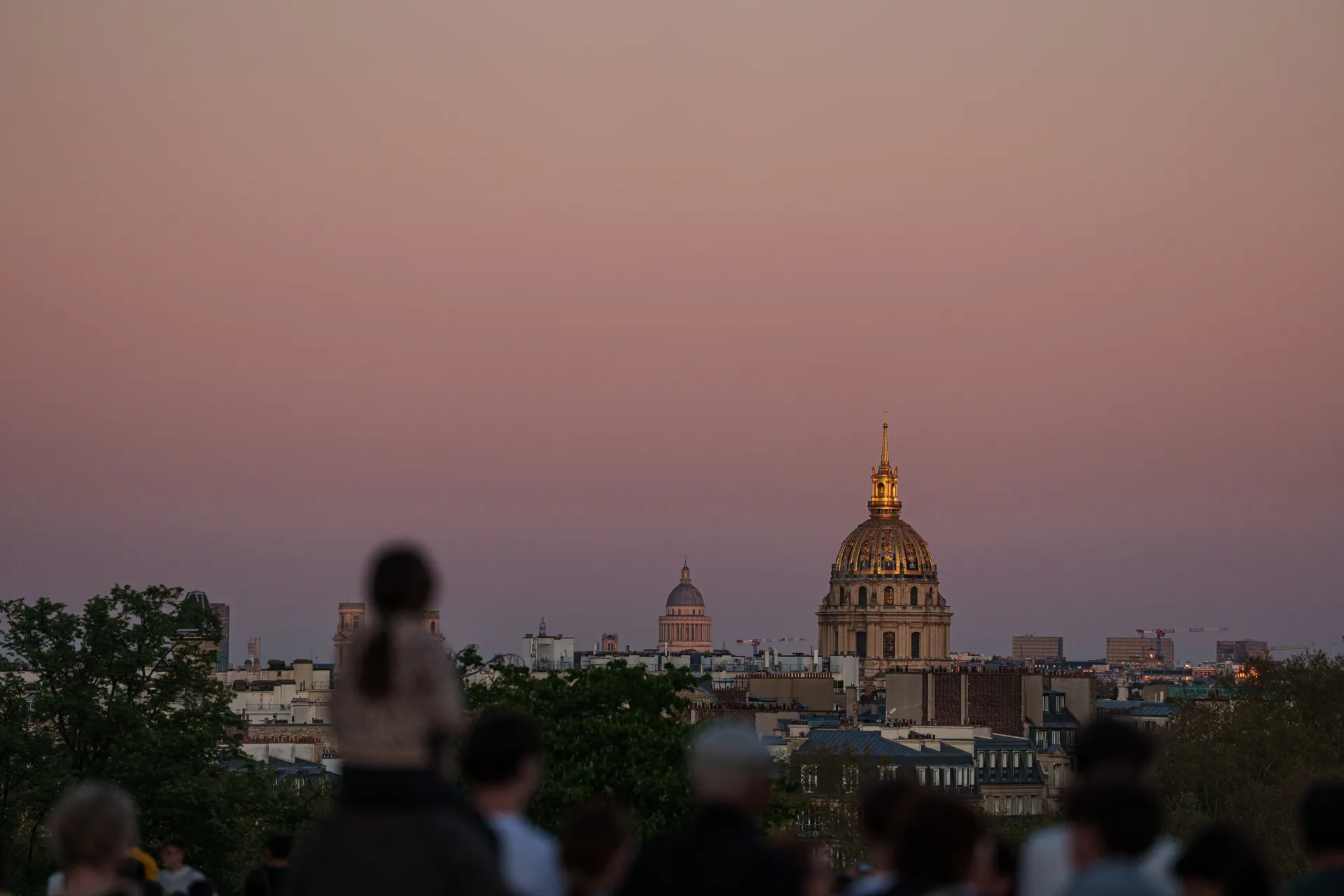 The golden dome of Les Invalides glows in pink dusk light above the Paris skyline, watched by silhouetted onlookers.