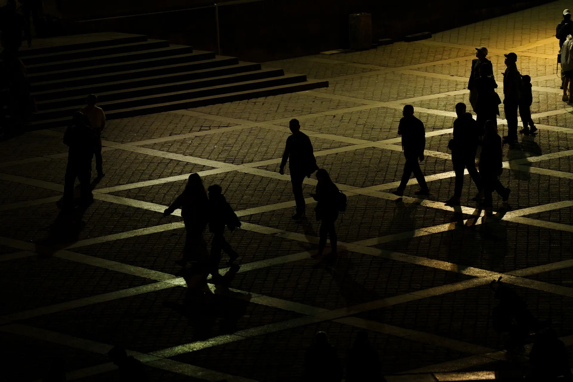 Silhouetted pedestrians cross a stone plaza at dusk, their long shadows stretching across diamond-pattern pavement.
