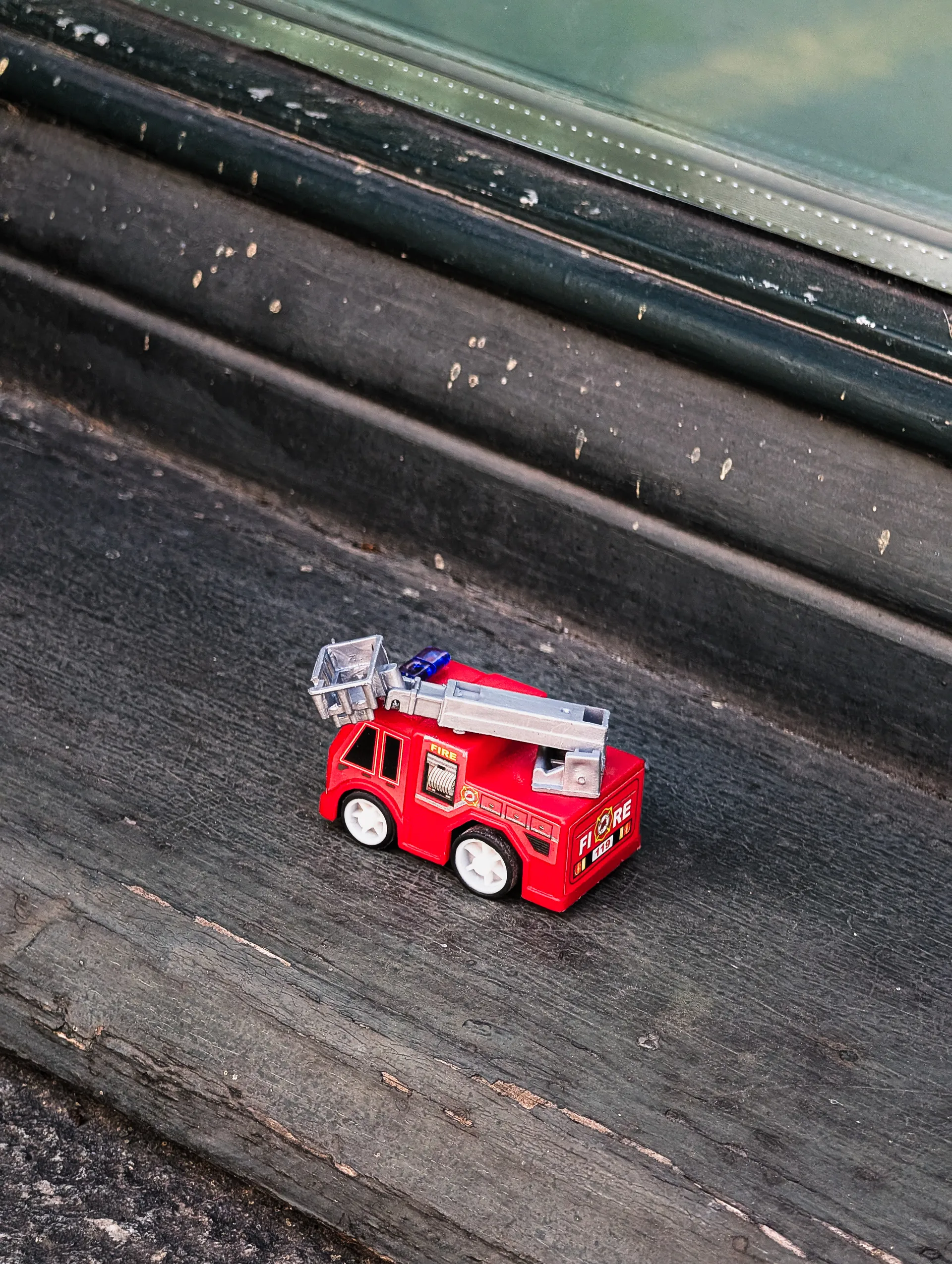 A miniature red fire truck sits on a weathered window ledge, marks of wear scattered across the dark surface.