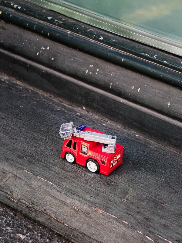 A miniature red fire truck sits on a weathered window ledge, marks of wear scattered across the dark surface.