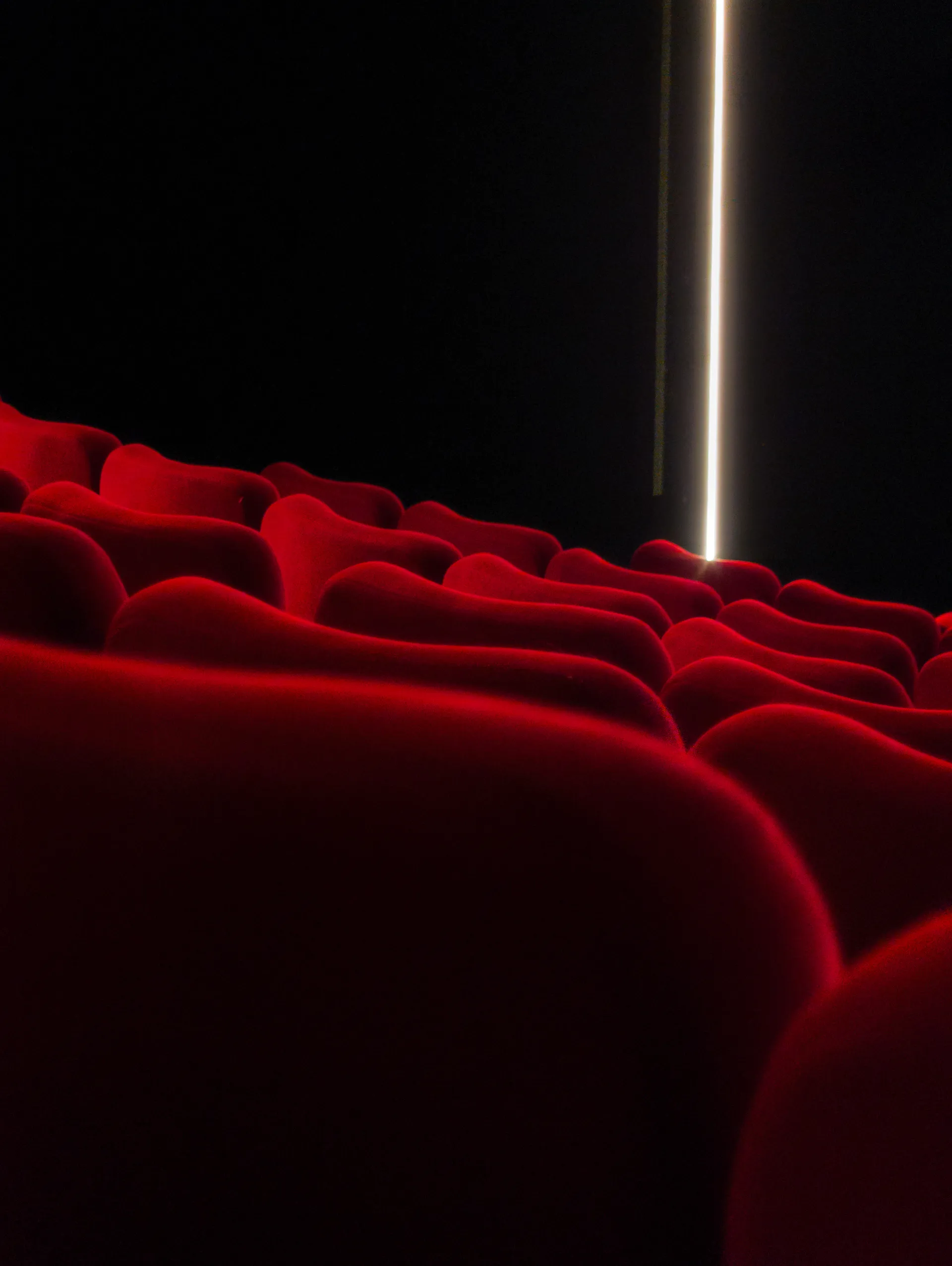 Rows of red velvet theater seats recede into darkness with a thin vertical strip of light between them.