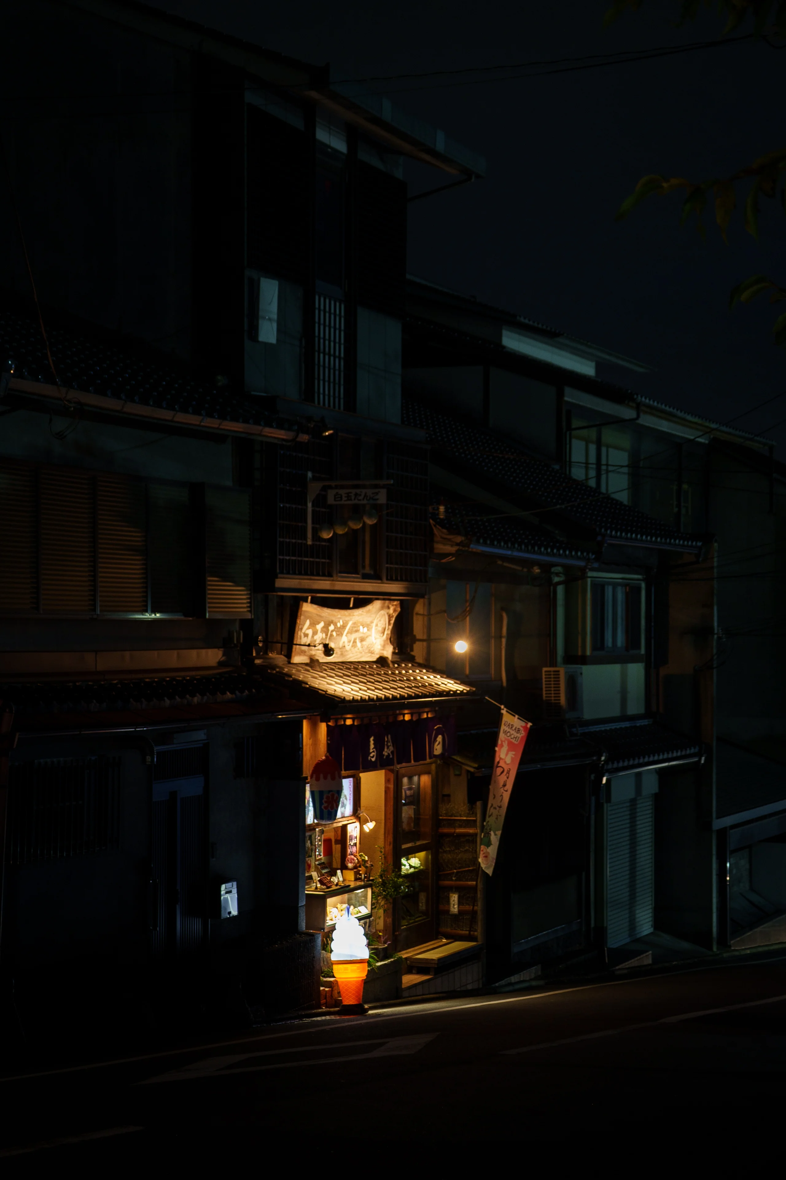 A small traditional shop glows warmly on a dark Kyoto street at night, its illuminated storefront the only light among the wooden machiya facades.