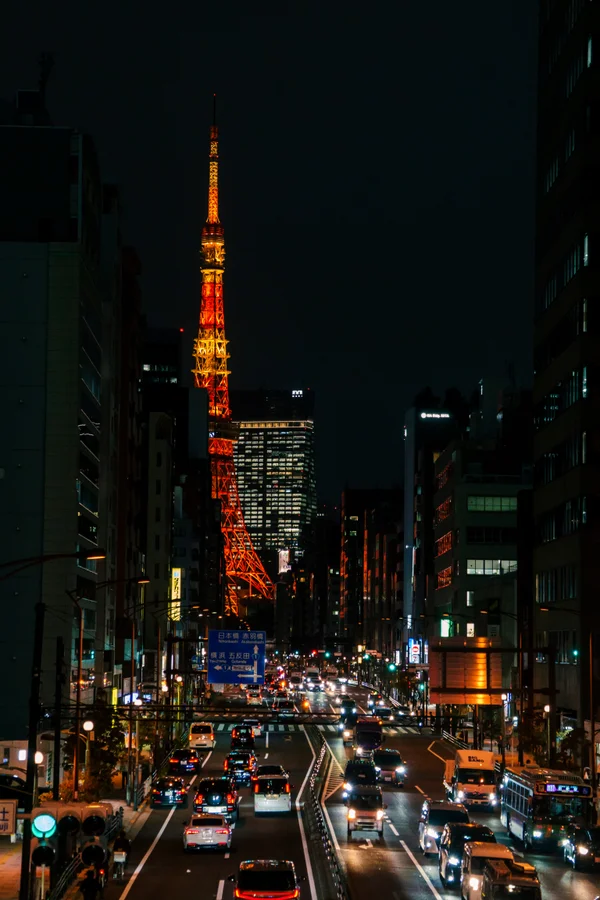 Tokyo Tower glows in red and gold above a busy night street, framed by silhouetted buildings as traffic flows past below.