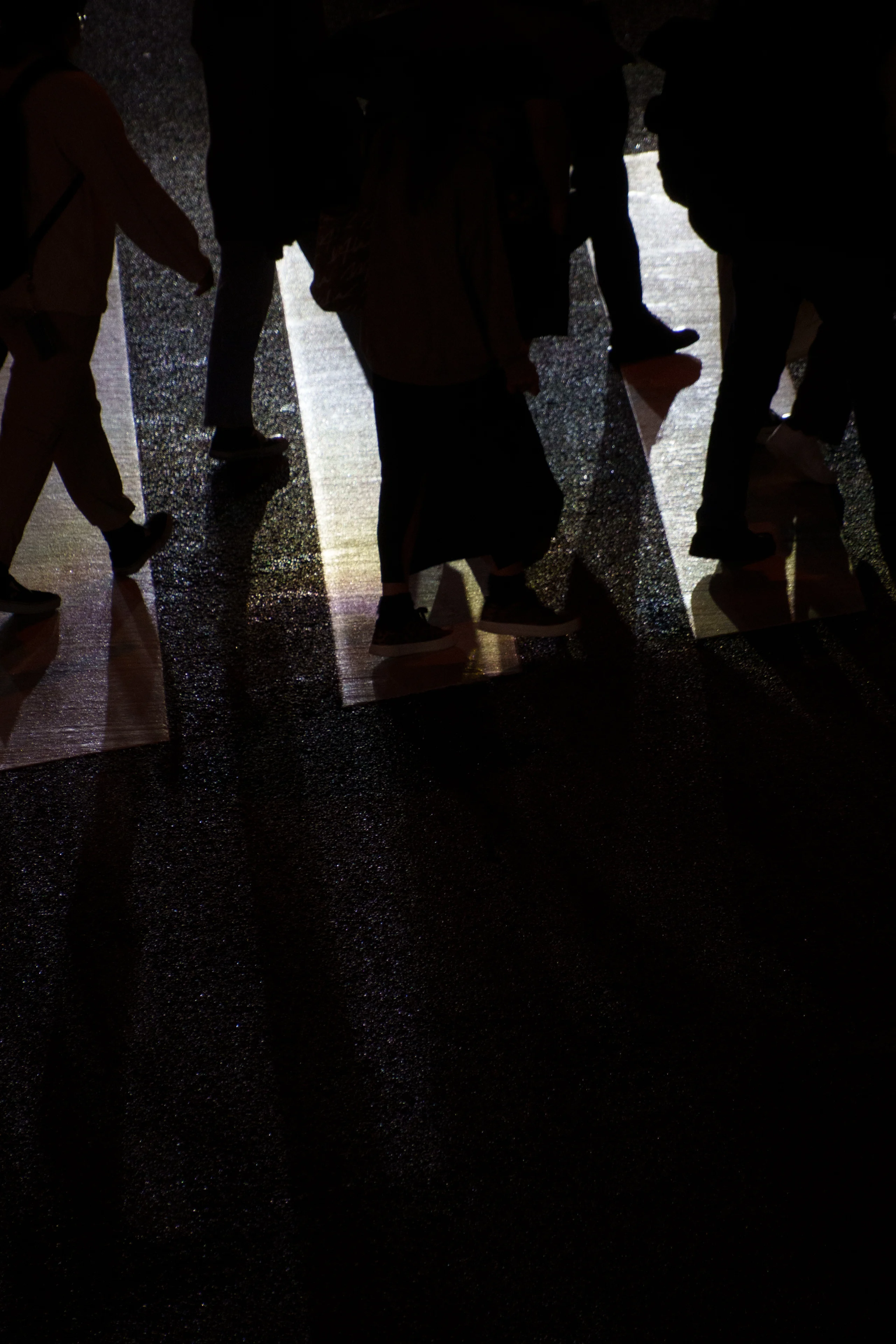 Silhouetted pedestrians cross a brightly lit crosswalk, their legs and long shadows cast across the dark, reflective asphalt.