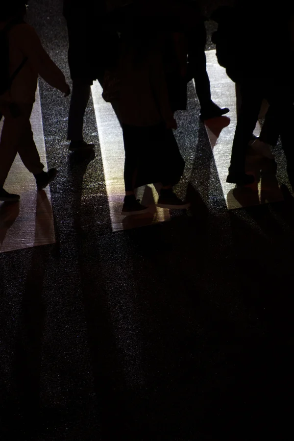 Silhouetted pedestrians cross a brightly lit crosswalk, their legs and long shadows cast across the dark, reflective asphalt.
