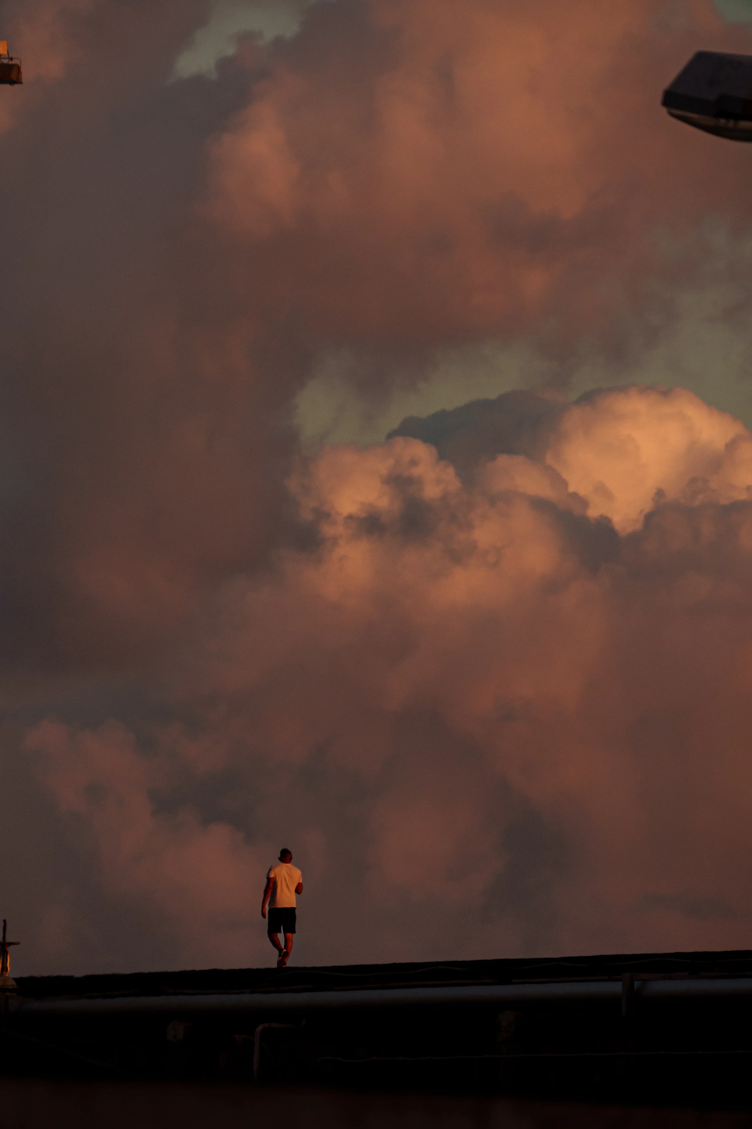 A lone figure walks along an elevated edge, silhouetted against towering sunset clouds glowing in warm orange and pink tones.