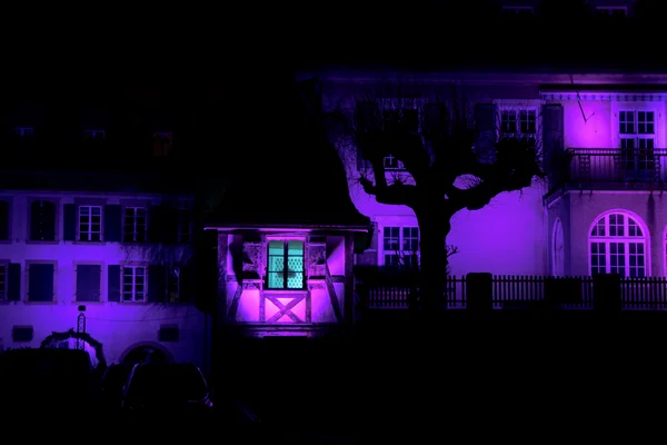 Traditional buildings bathed in violet light create a surreal nighttime scene, tree silhouettes adding depth.