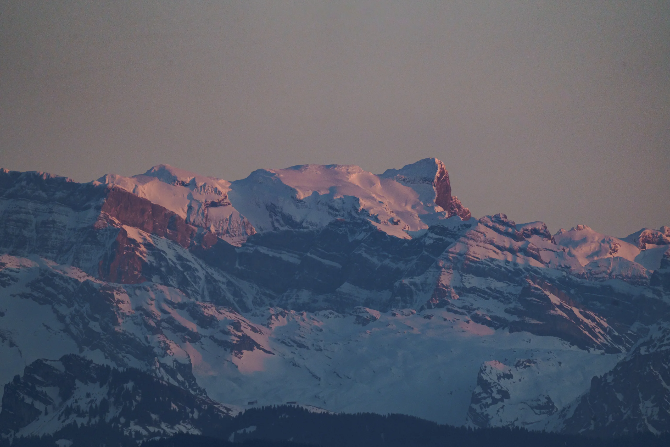 Snow-covered Alpine peaks catch the last warm light of the day, their ridges glowing pink and orange against a muted sky.