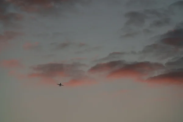 A lone airplane crosses a moody sky streaked with pink and grey clouds at dusk.
