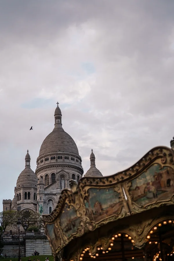 The domes of the Sacré-Cœur basilica rise into an overcast sky above the ornate, painted canopy of a vintage carousel.